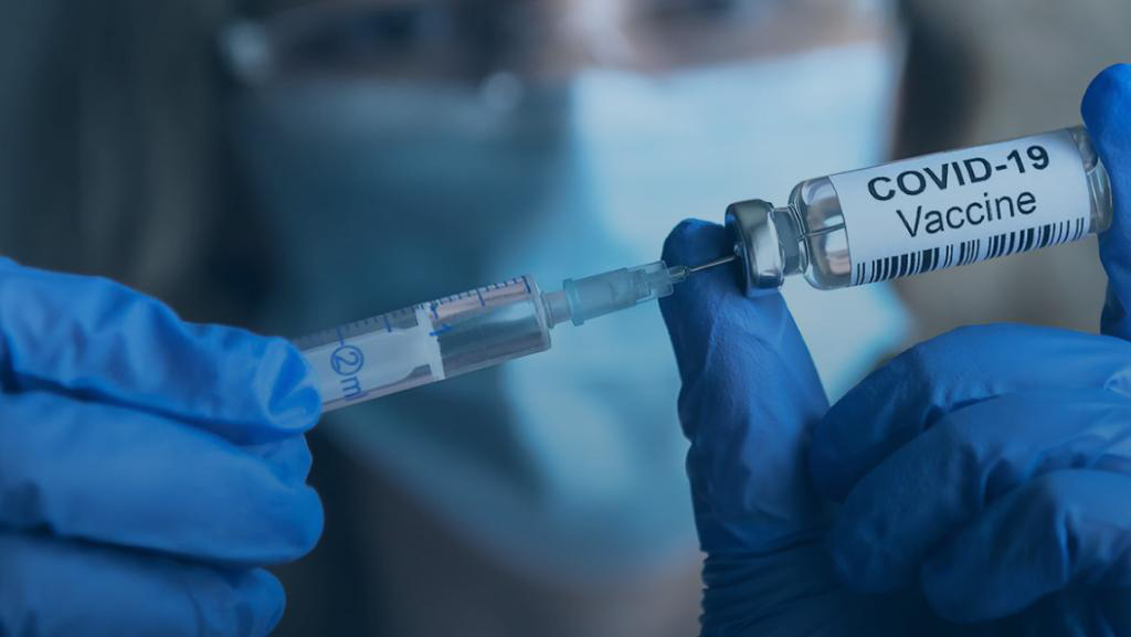 A person, wearing gloves and a surgical mask, handles a COVID-19 Vaccine vial and syringe.